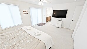 Une chambre à coucher avec un lit, une télévision, un ventilateur de plafond et une salle de bain visible à travers une porte ouverte.