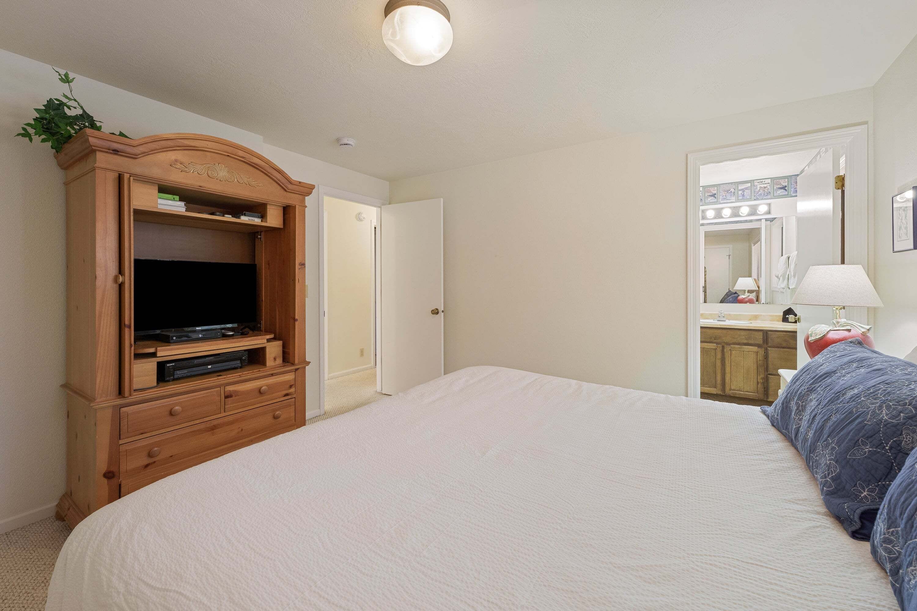 Une chambre à coucher avec un grand lit, une armoire en bois avec un téléviseur, et une salle de bain visible à travers une porte ouverte.