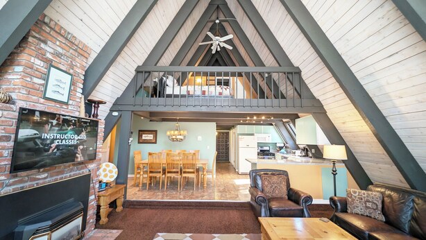 A spacious loft-style room with a high ceiling, exposed wooden beams, a fireplace, a dining area with wooden chairs, a kitchen in the background, and a balcony with a view. - Trapper 11 - A-Frame, Wood Stove, Pool Table (Sunriver)