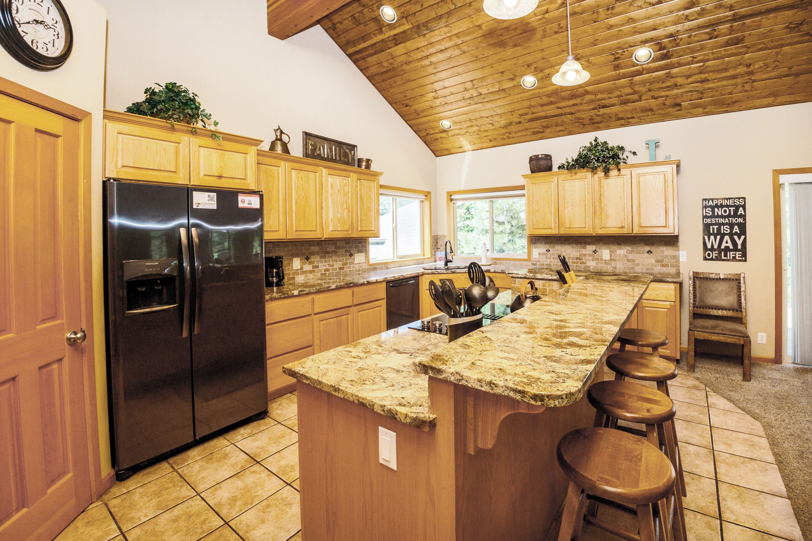 A kitchen with wooden cabinets, a granite countertop, a stainless steel refrigerator, and a bar stool area.
