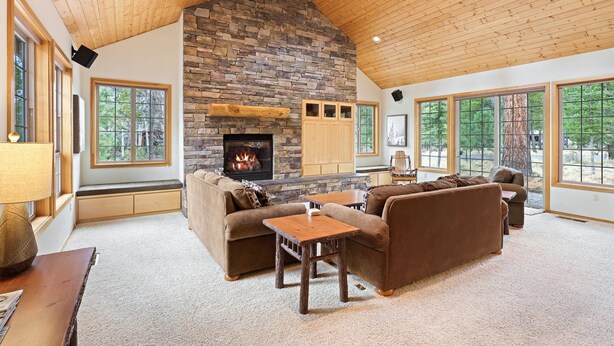 A spacious living room with a stone fireplace, wooden ceiling, and large windows offering a view of the outdoors. - Tokatee 14 - Beautiful & Spacious, SHARC, Ping Pong, A/C, Hot Tub (Sunriver)