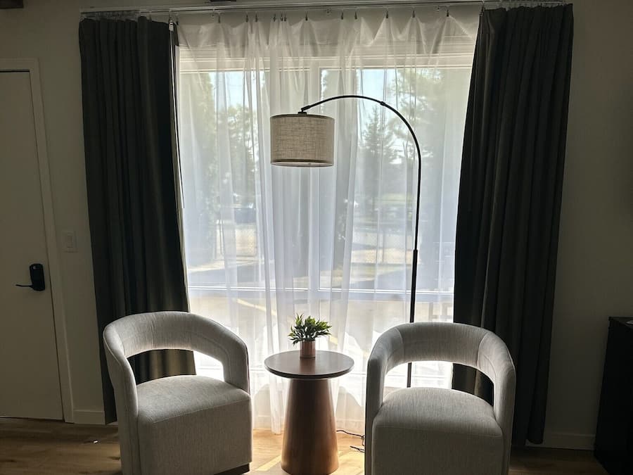 Et værelse med to lænestole, et lille bord med en plante, en gulvlampe og et stort vindue med lette gardiner.