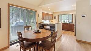 A dining area with a round wooden table and chairs, a kitchen with wooden cabinets, and a large window with blinds. - Summit View 5 - Updated Single Level, SHARC, A/C, Hot Tub (Sunriver)