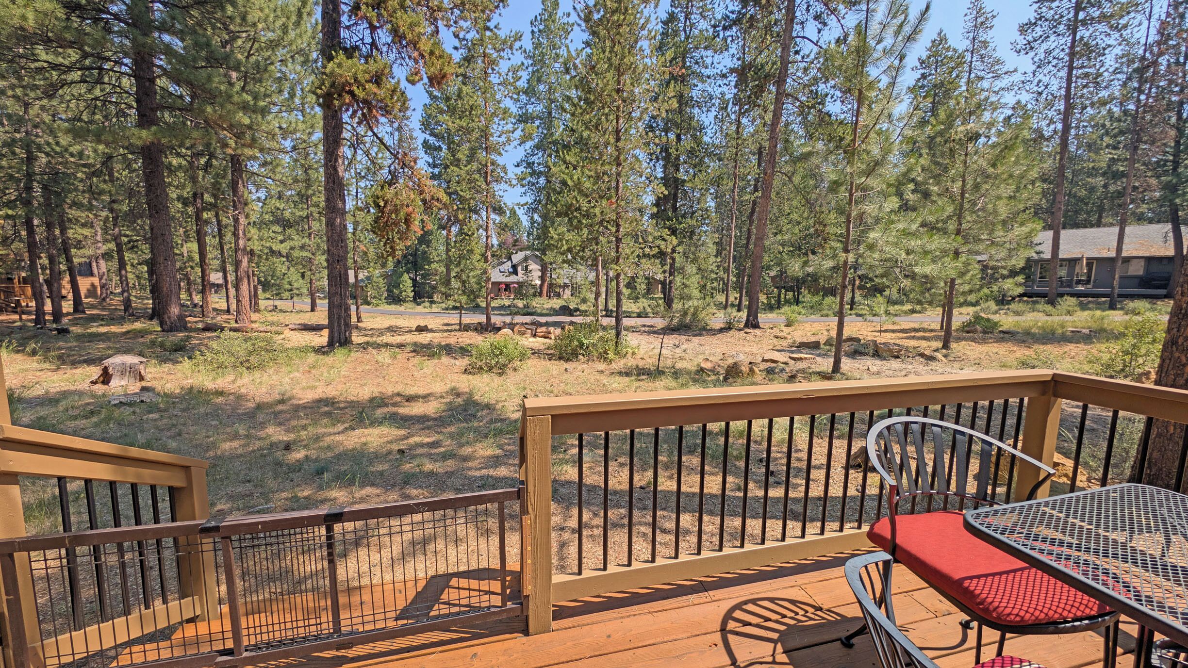 A wooden deck with a metal table and chairs, overlooking a forested area with tall trees and a clear sky.