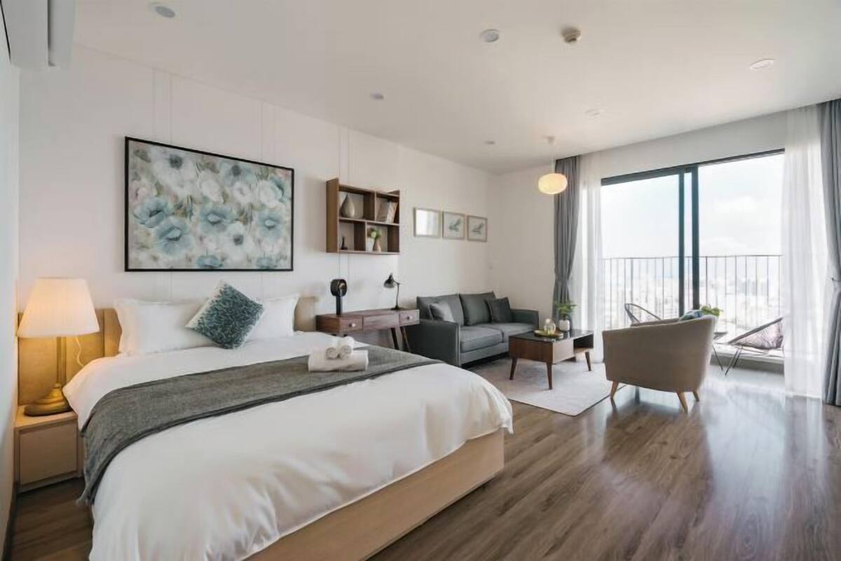 A modern hotel room with a large bed, a sofa, a chair, a balcony with a view, and a painting on the wall.