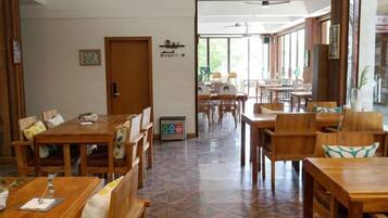 A dining area with wooden tables and chairs, a tiled floor, and a view through an open doorway to another room.