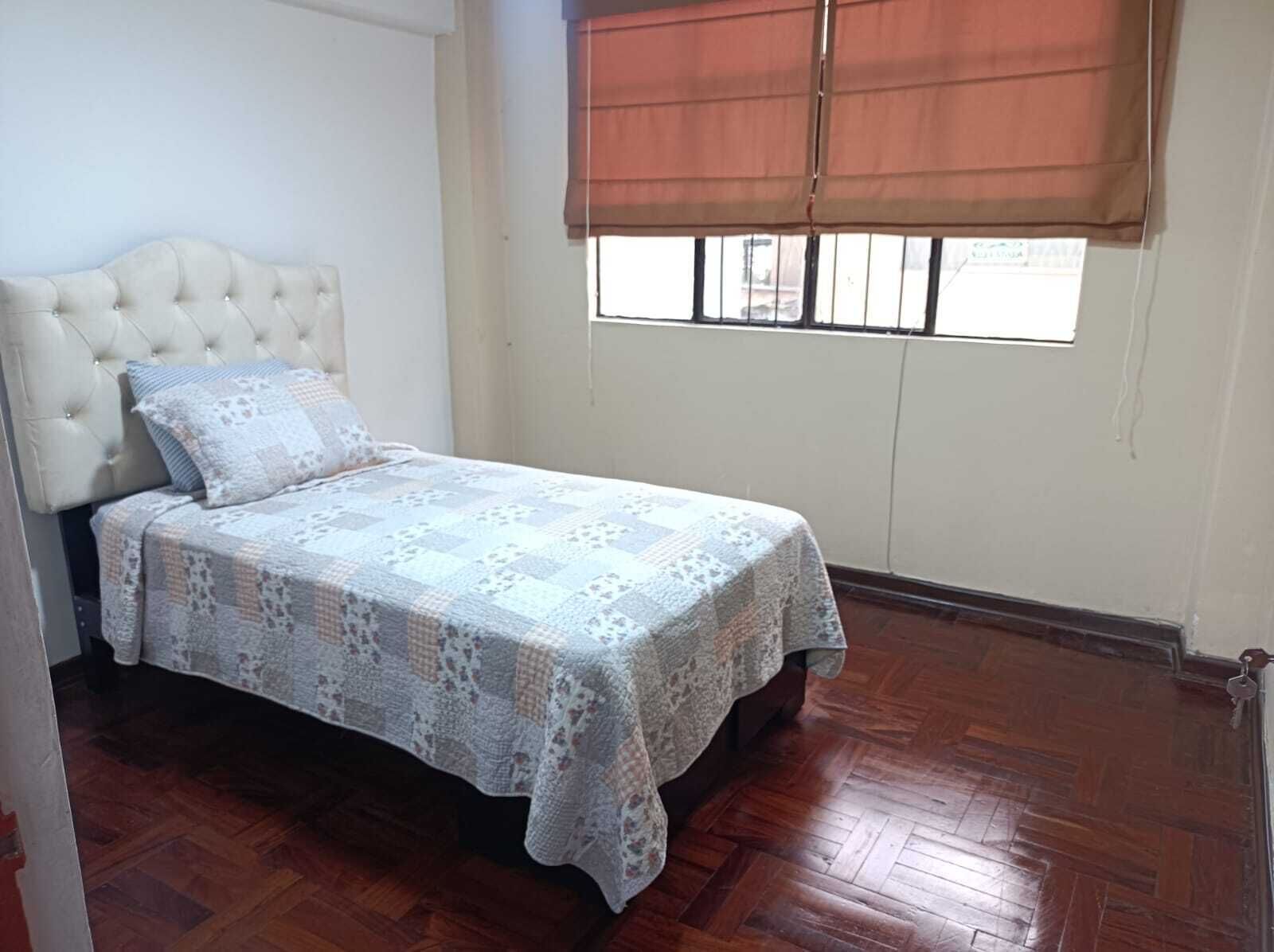 A bedroom with a tufted headboard, a quilted bedspread, and a window with a brown blind.