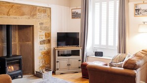 A cozy living room with a stone fireplace, a television on a wooden stand, a comfortable sofa, and a window with curtains. - Cottage in Grassington: Cosy 2-Bedroom Retreat (Grassington)