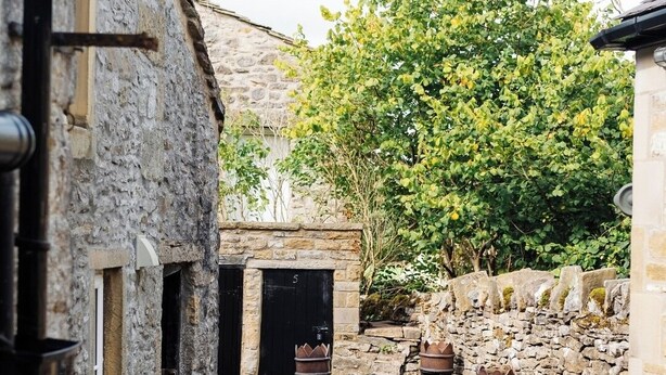 A narrow cobblestone alley with stone buildings, a tree-lined path, and outdoor seating with metal chairs and a table. - Cottage in Grassington: Cosy 2-Bedroom Retreat (Grassington)