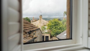 A view through a window of a stone building with a traditional roof, surrounded by greenery and a cloudy sky. - Cottage in Grassington: Cosy 2-Bedroom Retreat (Grassington)