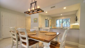 A dining area with a wooden table and white chairs, a chandelier, and a kitchen in the background.