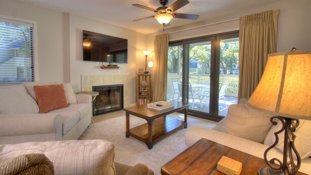 A living room with a fireplace, a ceiling fan, a television mounted on the wall, and a view of the outside deck through a glass door.