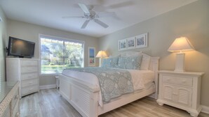 A bedroom with a bed, a ceiling fan, a TV, a window with blinds, and a nightstand with a lamp.
