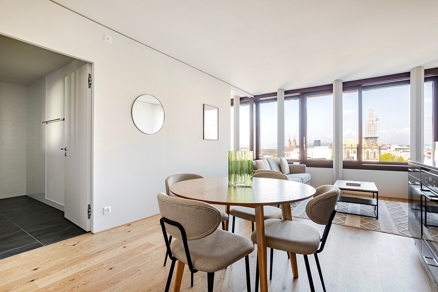 En moderne stue med et rundt spisebord, behagelige siddepladser, et stort vindue med byudsigt og en dør til et andet rum.