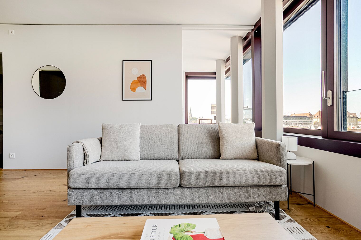 A modern living room with a grey sofa, a round mirror, and a framed abstract painting on the wall.