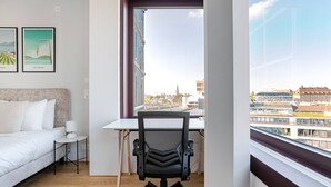 A bedroom with a bed, a desk, and a chair, a window with a city view, and two framed pictures on the wall. - Blueground | Basel-Vorstaedte, view, nr Kunstmuseum (Basel)