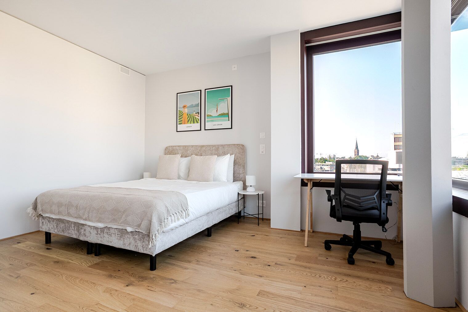 A bedroom with a bed, a desk with a chair, a window with a view of a cityscape, and two framed pictures on the wall.
