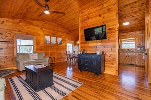 A cozy cabin interior with a wooden interior, a sofa, a coffee table, a TV mounted on the wall, and a kitchen area.