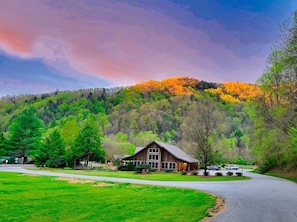 A rustic barn-style building with a large porch and picnic tables, surrounded by lush green fields and a forested hill in the background.
