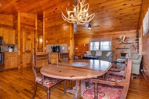 A wooden cabin interior with a dining table, chairs, a chandelier, a sofa, and a fireplace.