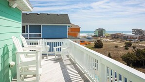 Eine Holzterrasse mit weißen Stühlen, ein blaues Gebäude und der Blick auf Strand und Meer.