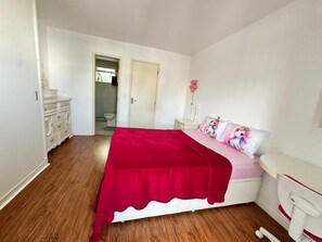 Quarto com piso de madeira, cama com cobertor vermelho, mesa de cabeceira com abajur rosa e cômoda branca.