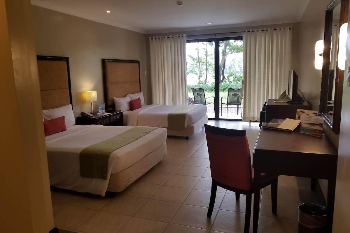 Habitación de hotel con dos camas, un escritorio, una silla, una lámpara y vistas al exterior a través de amplios ventanales.