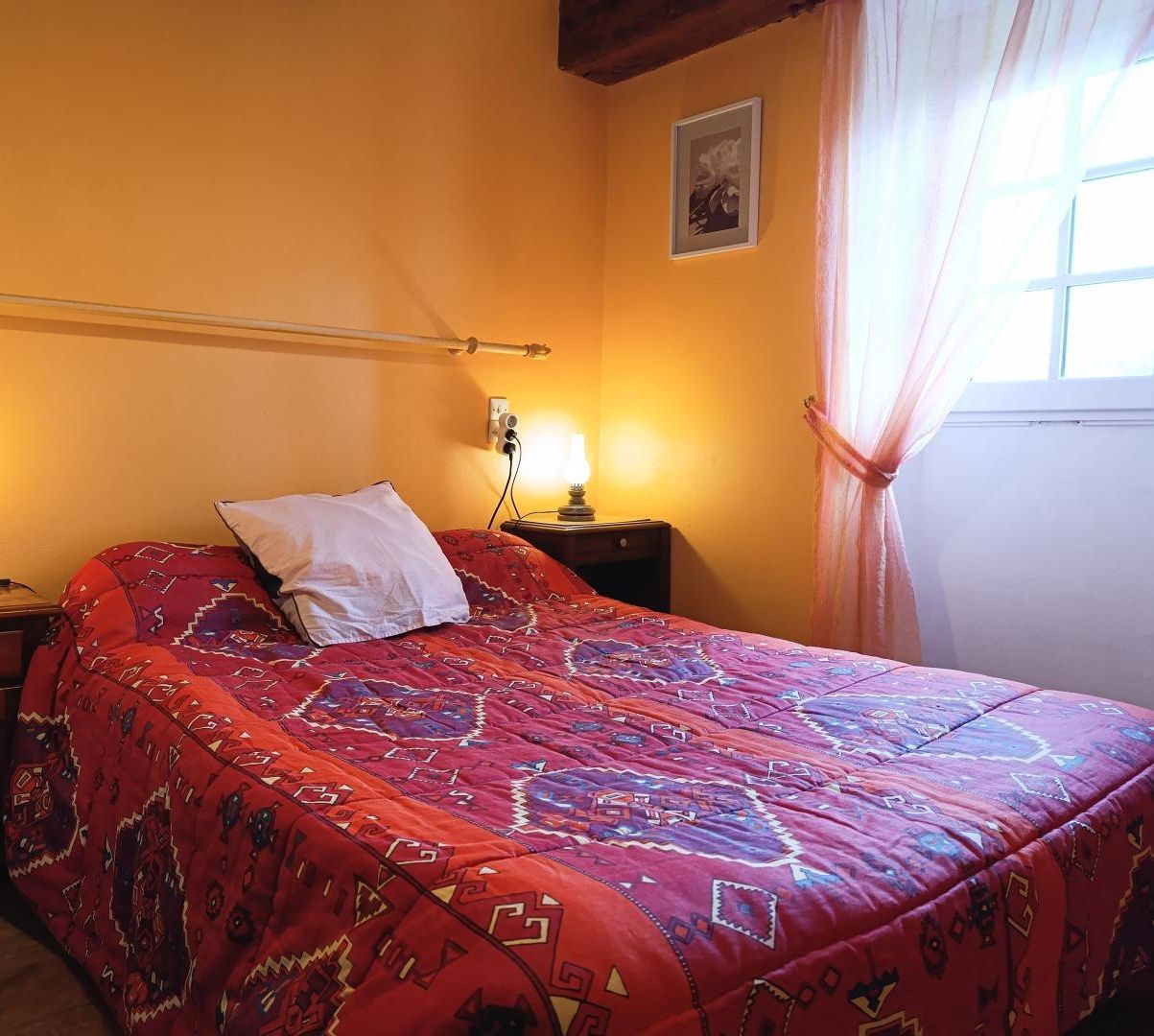 A bedroom with a bed, two bedside lamps, a nightstand, a window with curtains, and a framed picture on the wall.