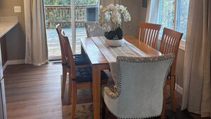 A dining area with a wooden table, chairs, and a chandelier. There is a view of a balcony with a railing and trees. - Relaxing-Retreat, Anchorage Getaway, Cozy & Newly Remodeled (Anchorage)