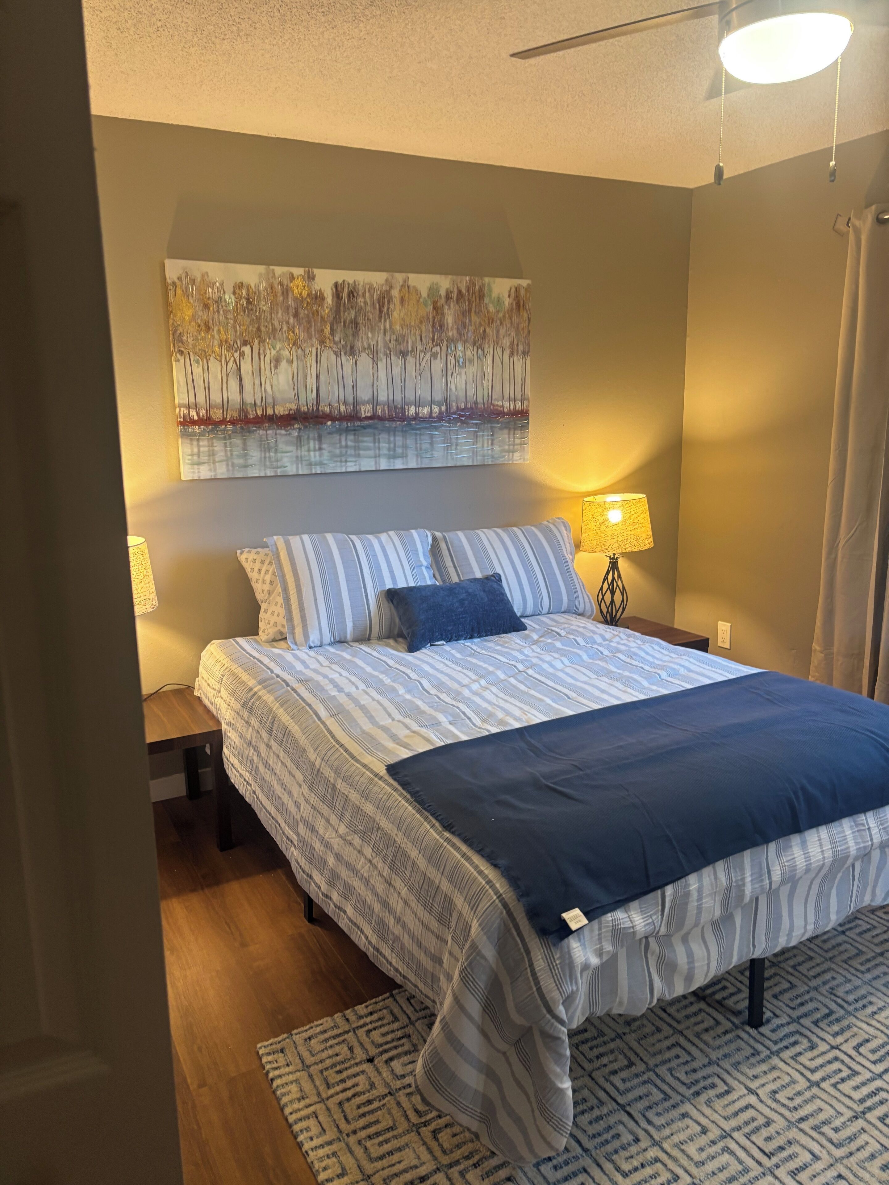 A bedroom with a bed, two bedside lamps, a painting on the wall, and a patterned rug.