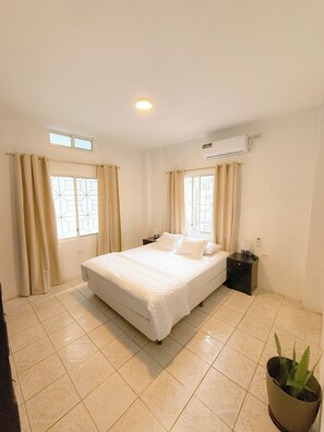 Een schone, goed verlichte slaapkamer met een bed, gordijnen, een nachtkastje, een airconditioning en een potplant.