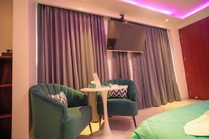 Una habitación con dos sillones verdes, una mesita blanca, una cama con ropa de cama verde y un ventanal con cortinas.