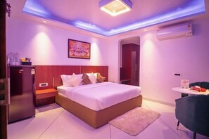 A modern hotel room with a bed, a small table, a chair, a refrigerator, and an air conditioning unit. - J60 Suites, Ashok Vihar - Near Netaji Subhash Place (North West Delhi)