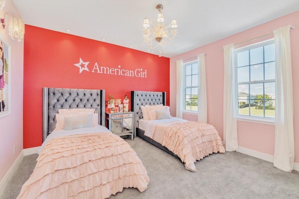 Ein Schlafzimmer mit zwei Betten, einer roten Wand mit 'American Girl'-Branding, einer Chandelüster und großen Fenstern.