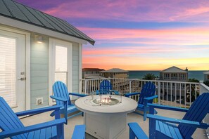 A balcony with blue Adirondack chairs, a fire pit, and a view of the ocean at sunset.