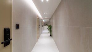 Un couloir d’hôtel moderne, avec un long passage rectiligne, des luminaires encastrés au plafond et un pot de fleurs placé sur le côté droit.