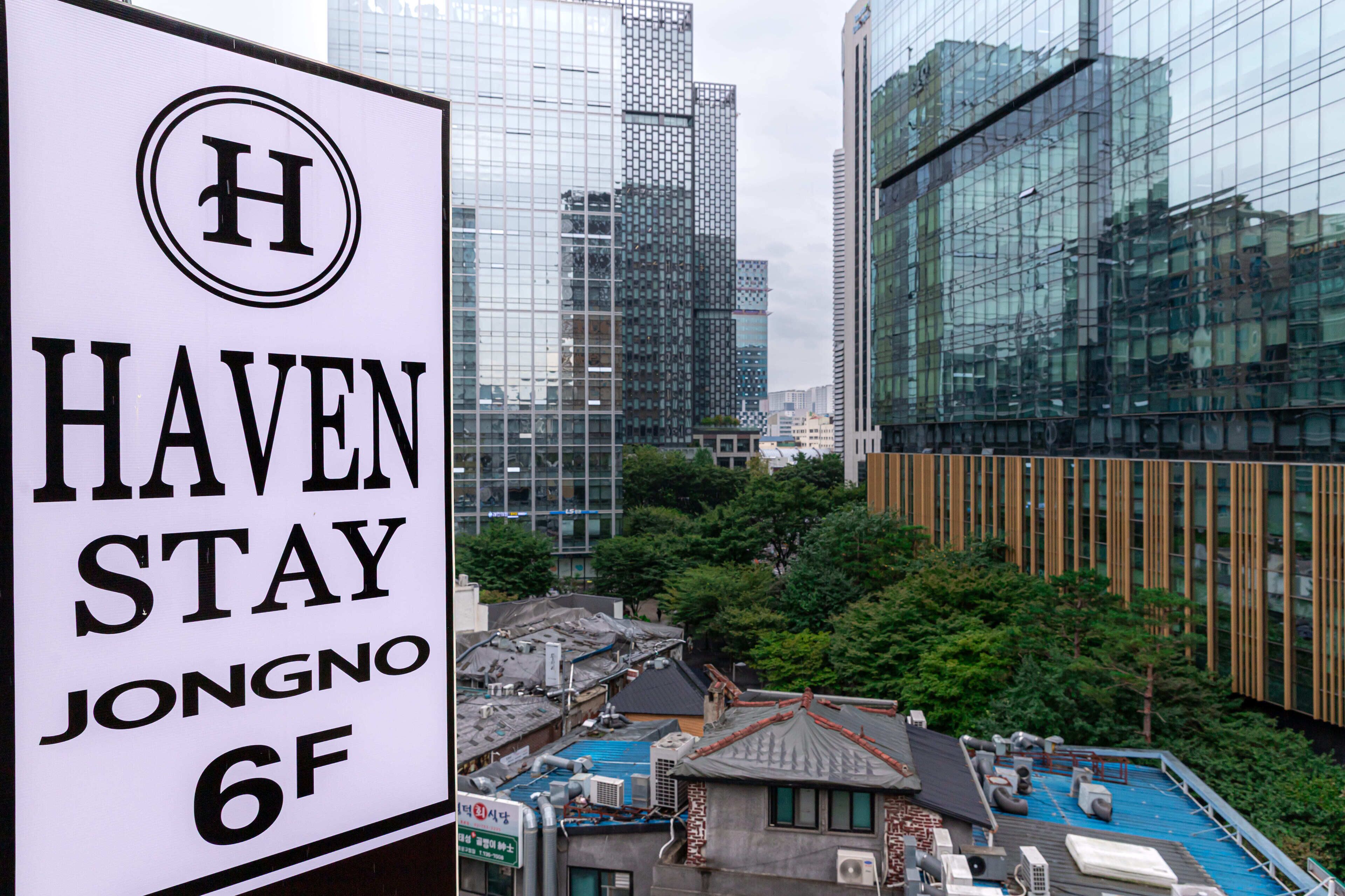 Señal de Haven Stay Jongno 6F con vista a edificios modernos y árboles al fondo.