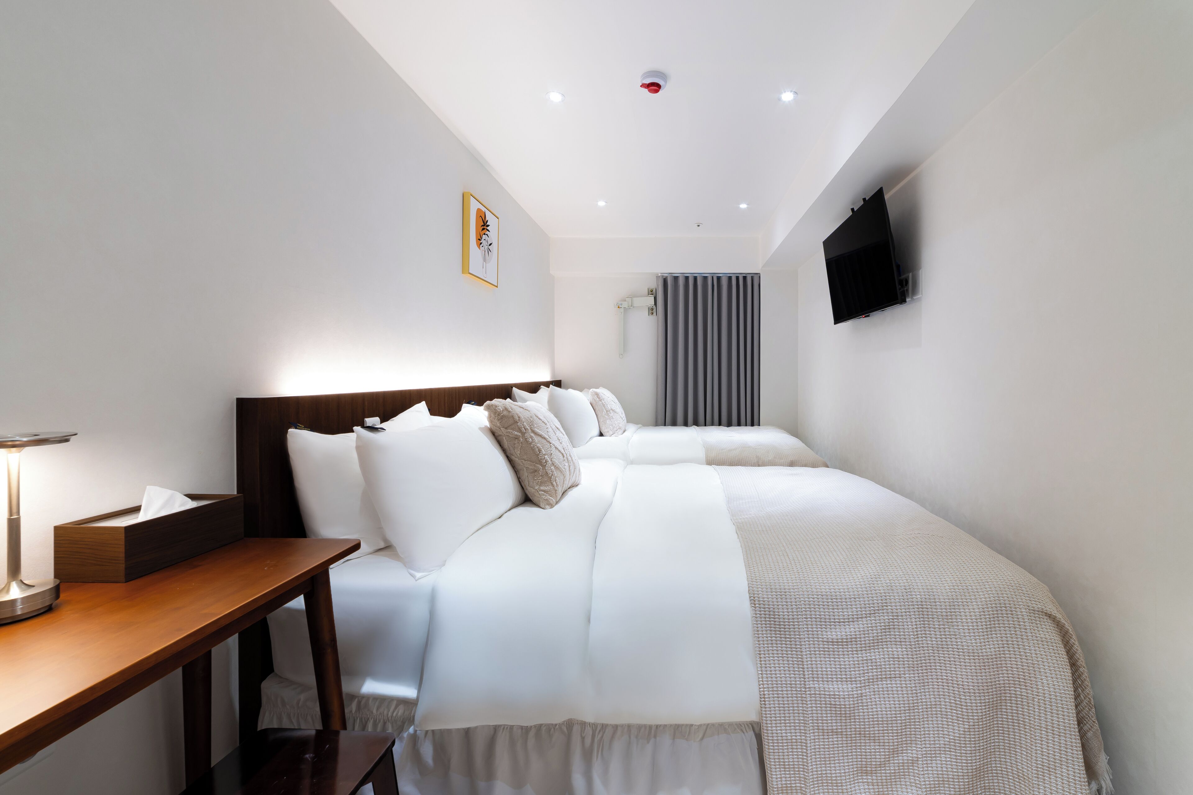 Une chambre d’hôtel avec deux lits, une table de chevet avec une lampe, un téléviseur fixé au mur et une fenêtre avec des rideaux.