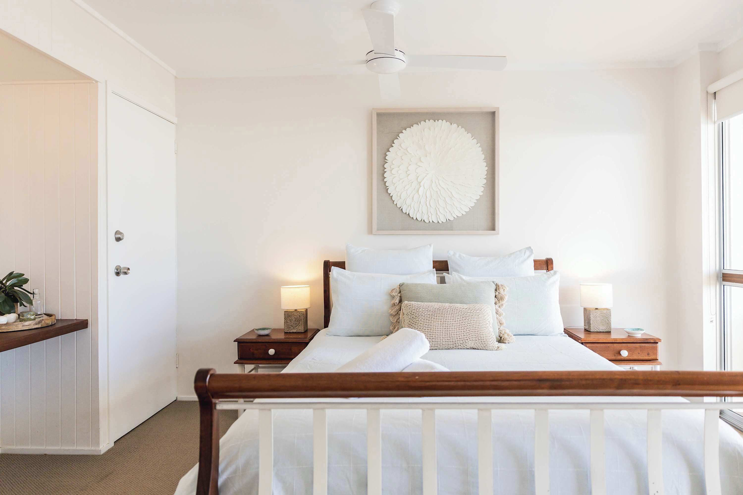 Ein Schlafzimmer mit einem hölzernen Bett, weißer Bettwäsche, einem Deckenventilator, Nachttischlampen und einem an der Wand befestigten Kunstwerk.