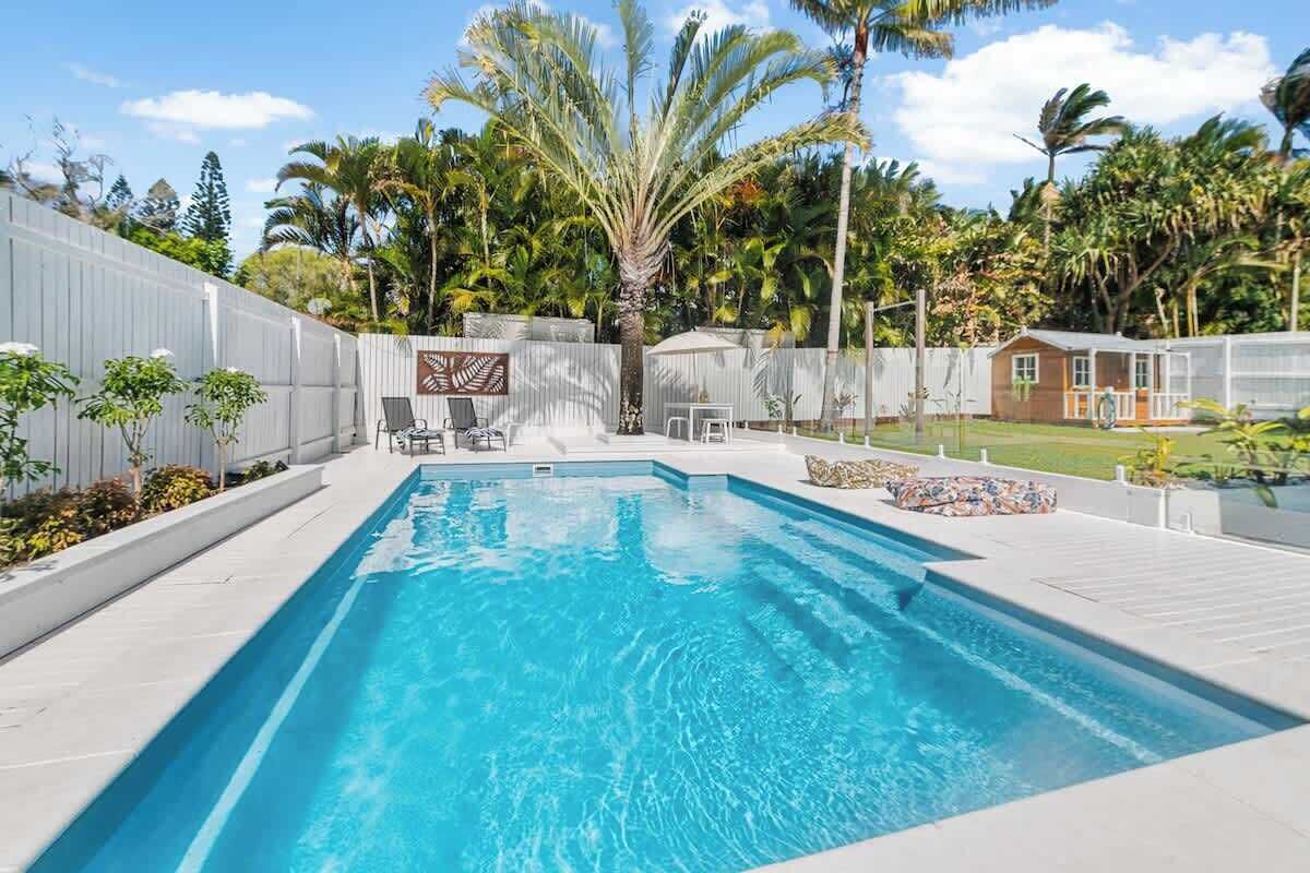 Une piscine rectangulaire dotée d’une plage blanche, entourée d’une clôture blanche et de palmiers.