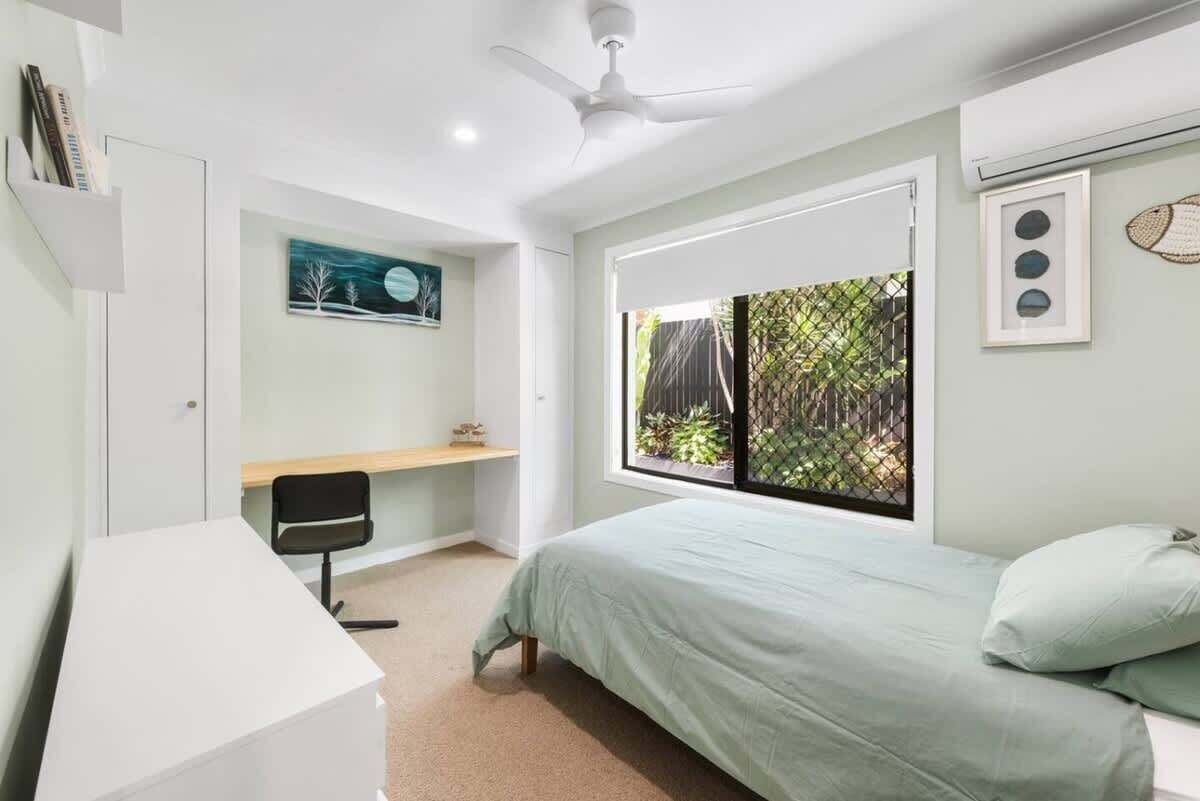 Een slaapkamer met een bed, een bureau, een stoel, een plafondventilator en een raam met uitzicht op groen.