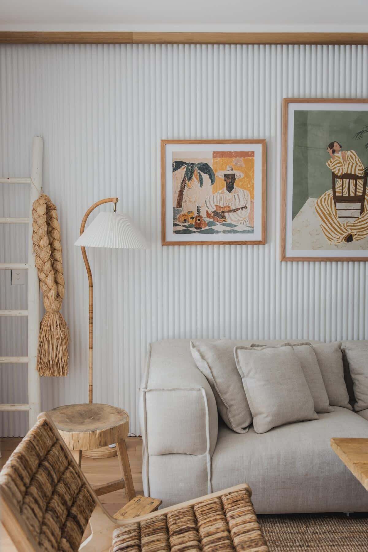 Een woonkamer met een lichtgrijze bank, een houten salontafel, een geweven stoel, een vloerlamp en twee kunstwerken in lijsten aan de muur.