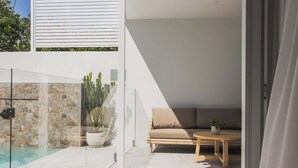 Un patio moderno al aire libre con piscina, un área de estar con sofá y mesa, y una pared de vidrio con una planta de cactus.