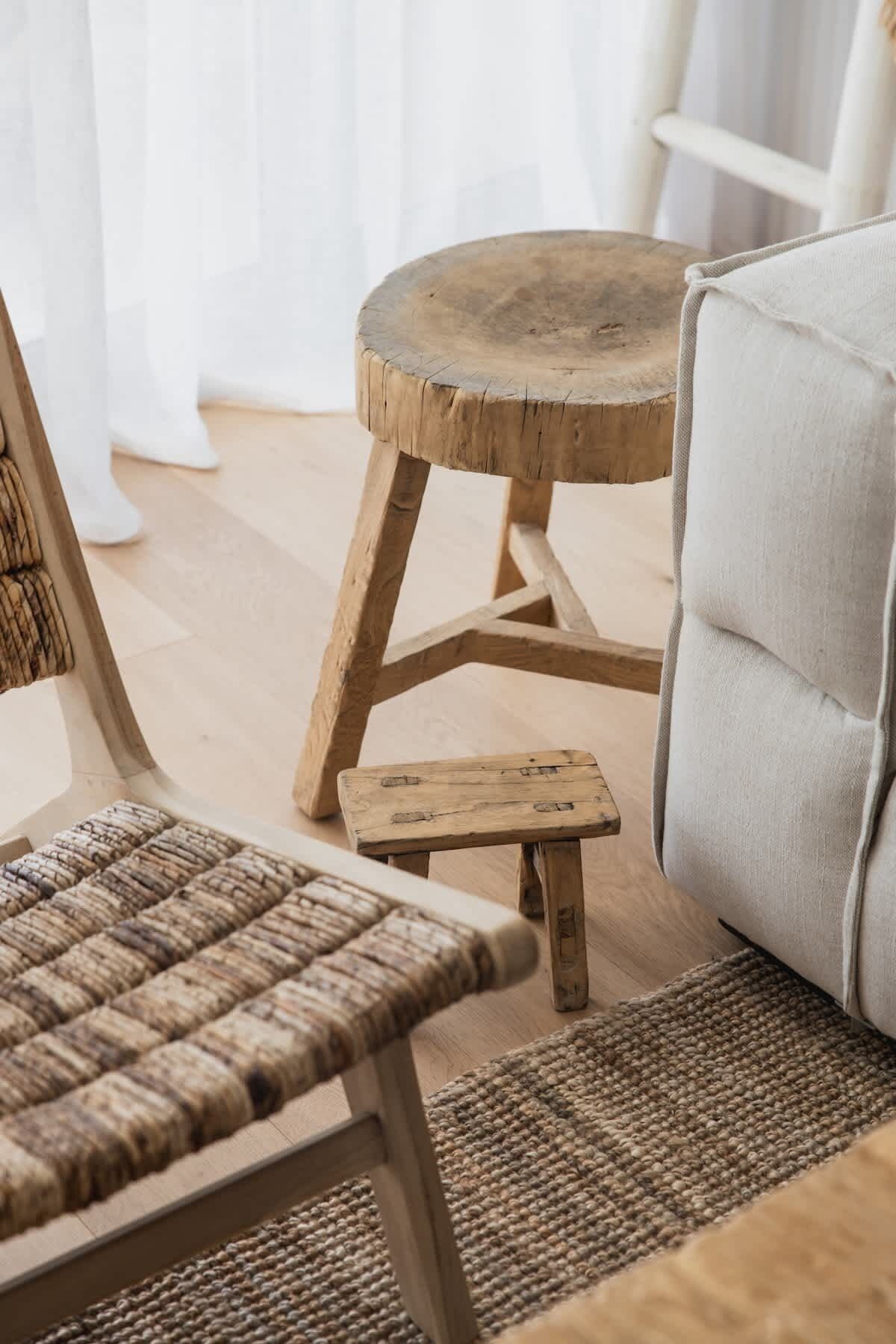 A rustic wooden stool with a woven seat and a matching small stool, placed on a woven rug in a room with a light-colored sofa and a chair with a woven backrest.