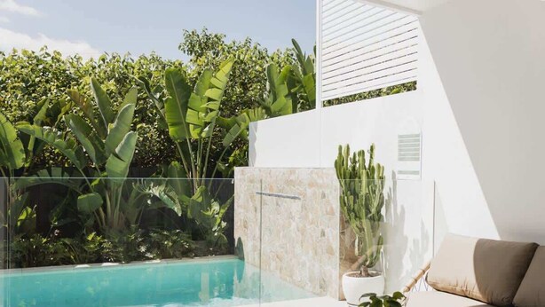 Zona de piscina exterior moderna con cerramiento de cristal, rodeada de exuberante vegetación tropical y un edificio blanco.