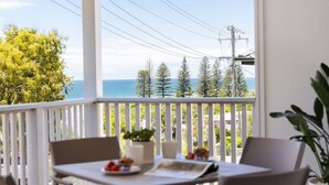 A balcony with a table and chairs, overlooking a coastal area with trees and power lines.