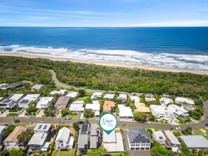 An aerial view of a coastal residential area with houses, a beach, and a logo for Noosa Holidays.