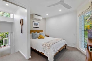 A bedroom with a bed, a ceiling fan, an air conditioning unit, a window with a view, and a small bedside table.