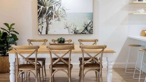 A dining area with a wooden table and chairs, a painting of a beach scene, and a potted plant. - Steps to Noosa River - a charming retreat in Noosa (Noosaville)