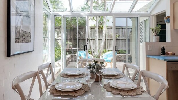 A dining area with a glass table set for four, surrounded by white chairs, with a view of a backyard through a glass door. - Cute Little Cove gem…walking distance to beach! (Noosa Heads)